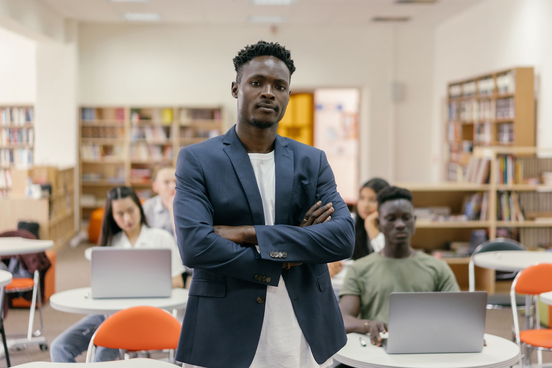 A confident Black student standing in a library with classmates studying at laptops in the background.